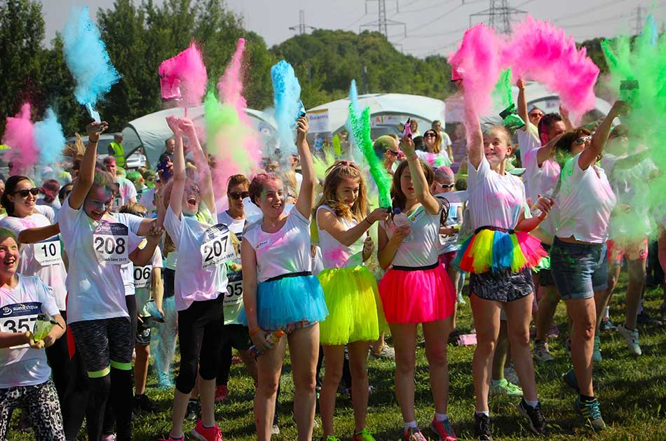 A group of people in colourful skirts and white shirts throw vibrant pink, blue, and green powder into the air during an outdoor event, smiling and celebrating on a sunny day.