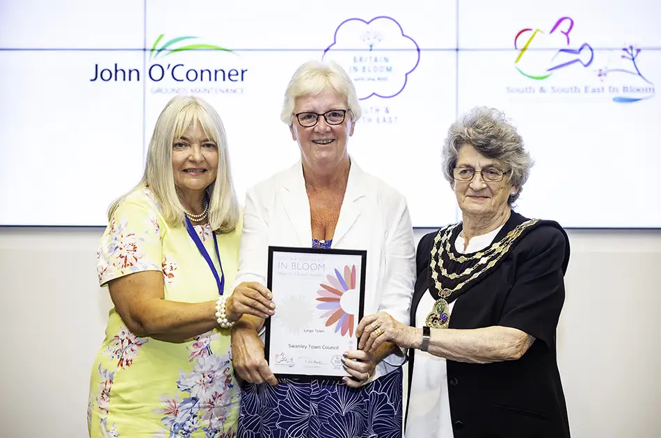 From left to right: The first lady is Cllr Lesley Dyball has long blonde hair and is wearing a yellow floral dress. The middle lady has short blonde hair and is wearing a white blazer over a blue patterned top. The lady on the right is Mayor of Swanley, Cllr Rita Wood, has short gray hair and is wearing a black outfit with a chain of office around her neck. They are standing in front of a white background with some logos or text visible. One logo says "John O'Conner" and there's another colorful logo visible as well. The certificate they're holding appears to say "In Bloom" on it.