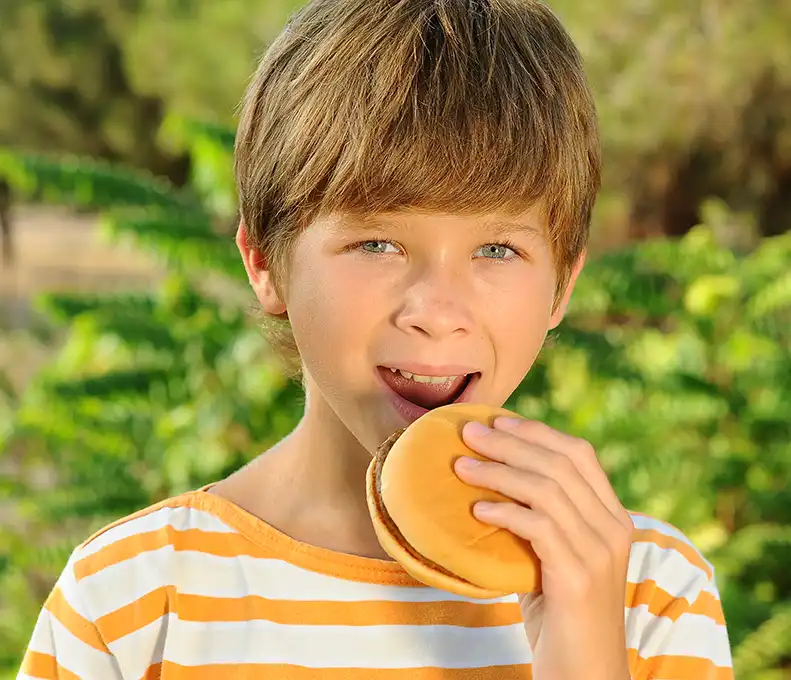 A young boy in a yellow and white striped shirt smiles whilst holding a burger outdoors, with green foliage in the background.