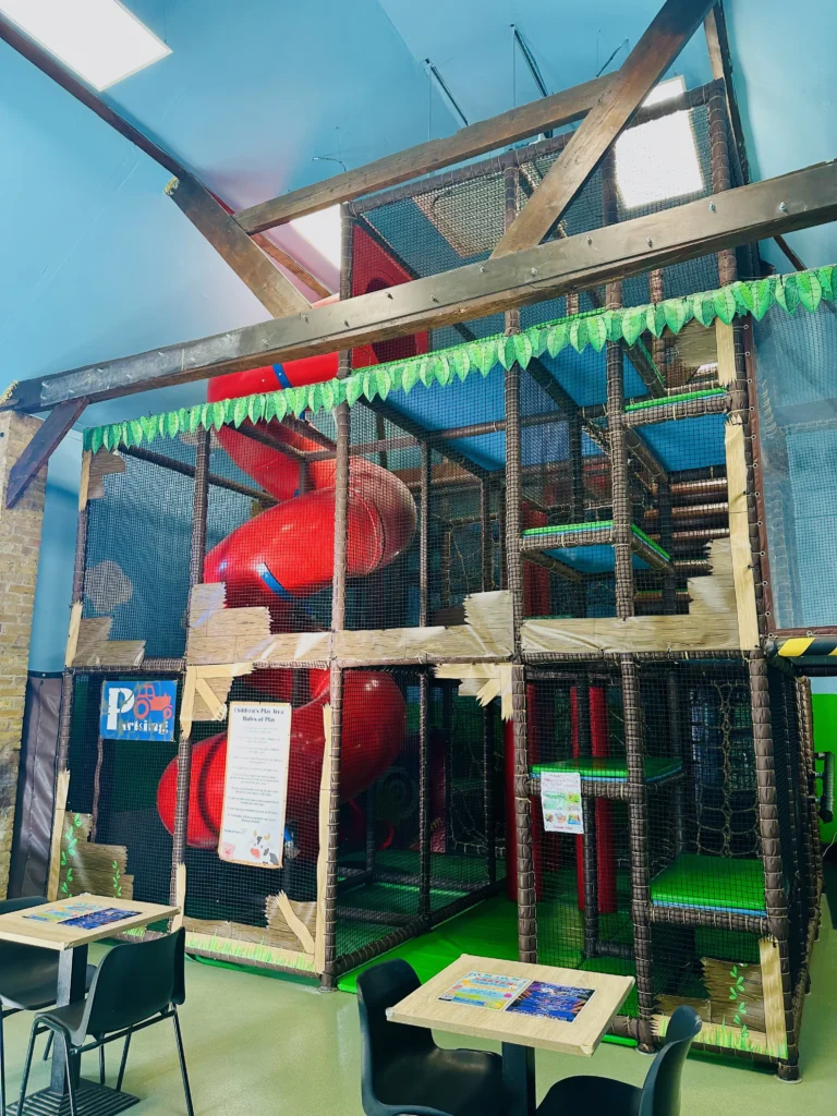 Indoor play area with a large climbing frame, red spiral slide, green padded floors, and netting. The structure is decorated with paper leaves, and there are tables and chairs in the foreground for seating.