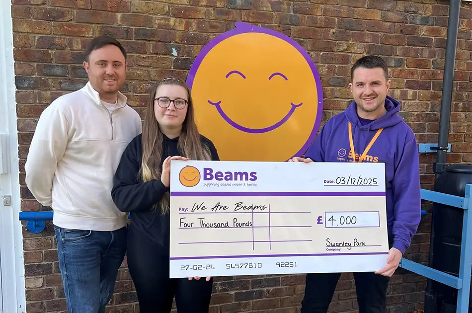 Three people stand in front of a brick wall, holding a large cheque for £4,000 to We Are Beams from Swanley Park, with a purple and orange Beams logo featuring a smiling face behind them.