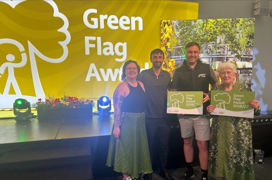 Four people stand on a stage, two holding Green Flag Award certificates, with a large “Green Flag Awards” sign and potted flowers in the background. All are smiling and dressed in smart-casual attire.