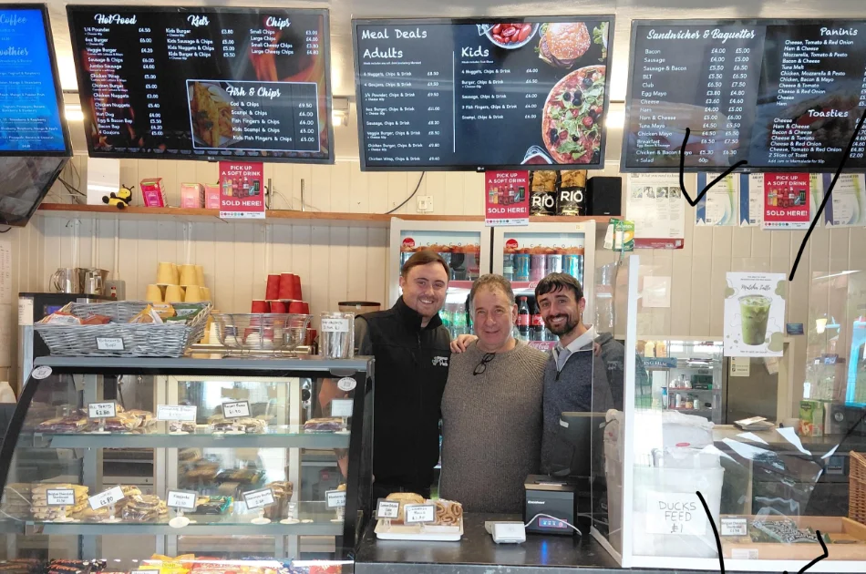 Three men stand behind the counter of a cafรฉ with menu boards displaying food options and prices on the wall behind them.